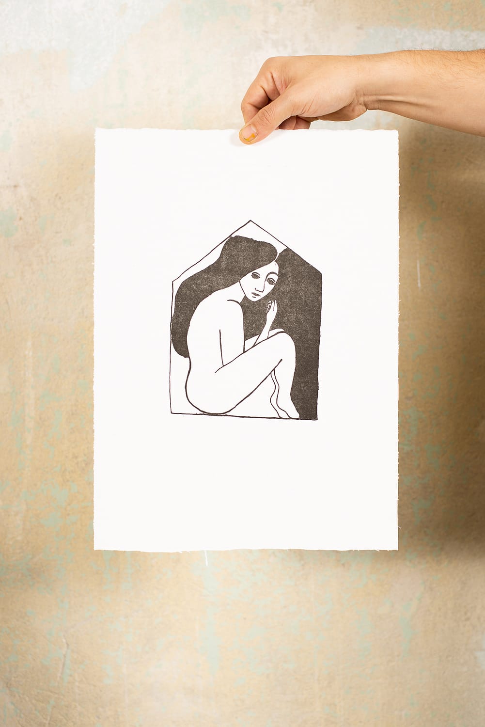 Hand hält lithographische Druckgraphik vor einer Wand. Auf der Graphik ist eine Frau zu sehen, die eine Form geduckt ist. Die Form erinnert an ein Haus und soll ein Gefühl des »eingesperrt seins« vermitteln.