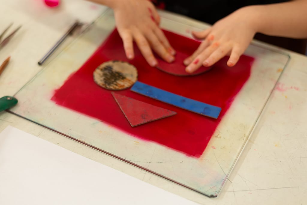 Children's hands apply stencils on glass plate for monotype