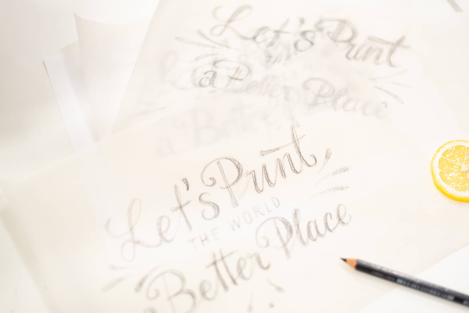 Transparentpapiere mit verschiedenen Schriftentwürfen, die den Schriftzug »Let’s Print the World a Better Place« zeigen. Daneben liegt ein Schreibwerkzeug und eine Zitronenscheibe.