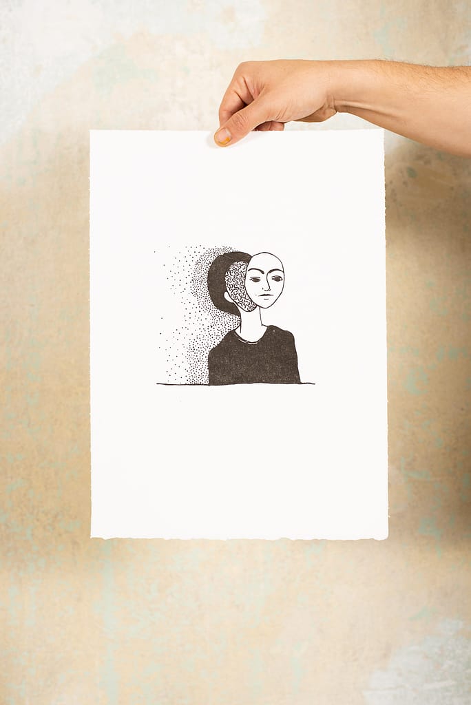 Hand hält lithographiscen Druckgraphik vor einer Wand. Auf die Graphik ist eine Illustration einer Frau mit ausgeschnittene Gesicht