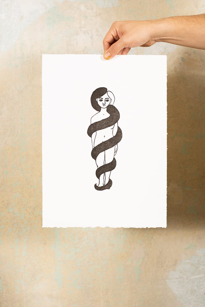 Hand hält lithographische Druckgraphik vor einer Wand. Auf der Graphik ist eine Frau zu sehen, der Haar sich in einem dicken Zopf um sie windet. Der Opfer erinnert an eine Schlange und die gesamte Figur damit an das Schlangensymbol für Gesundheit.