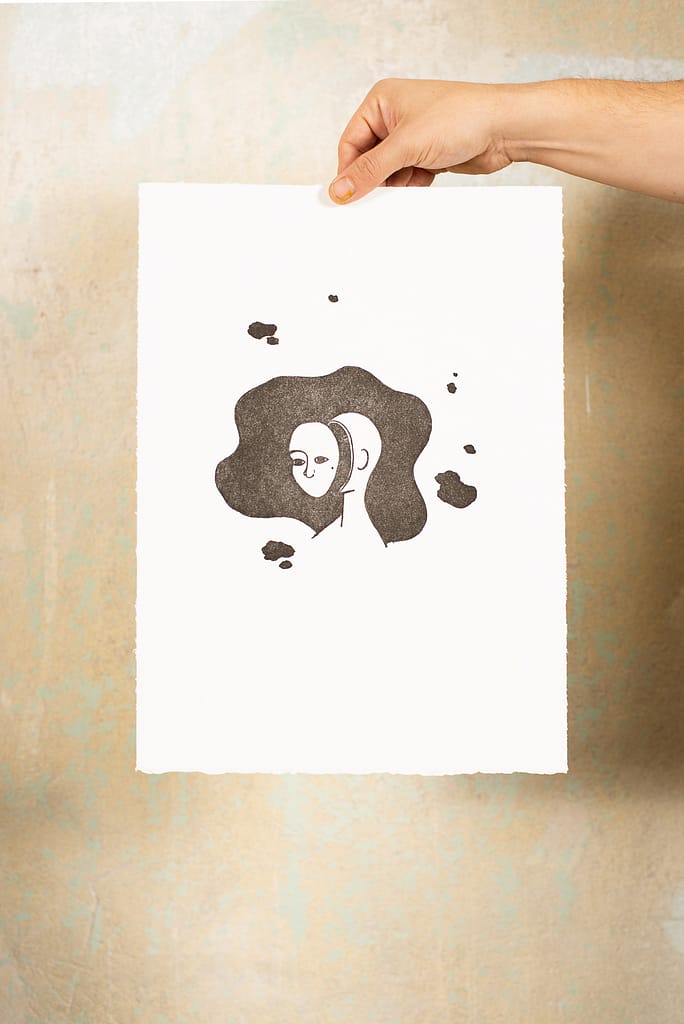 Hand hält lithographiscen Druckgraphik vor einer Wand. Auf die Graphik ist eine Illustration einer Frau mit ausgeschnittene Gesicht vor einem schwarzen Fleck